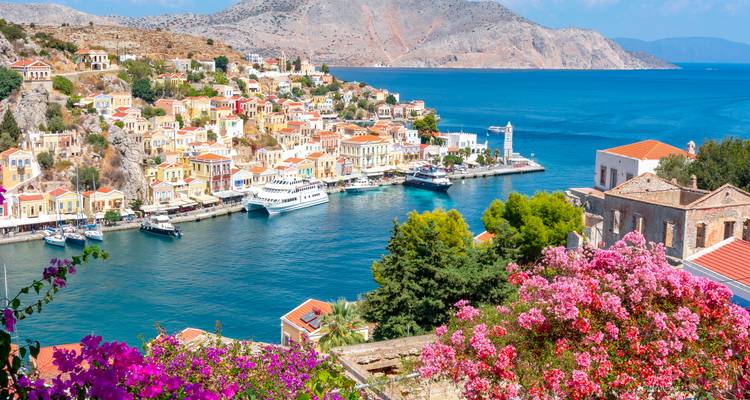 Village coloré à flanc de colline avec des maisons pastel et des yachts encadrant une baie turquoise bordée de bougainvilliers roses éclatants.