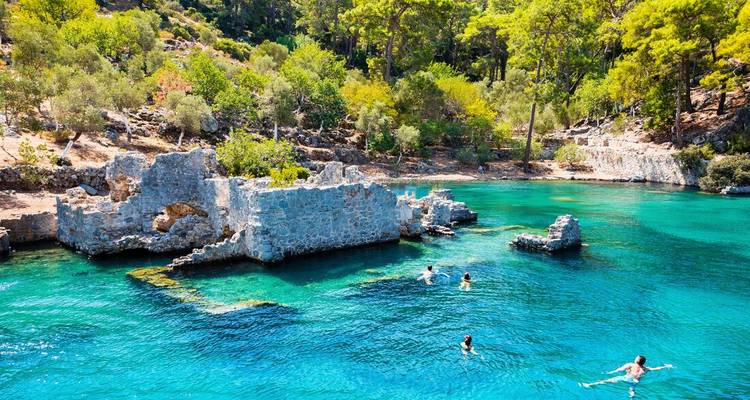 Swimmers explore turquoise waters around submerged ruins and pine-covered shore in Turkey’s Göcek Bay