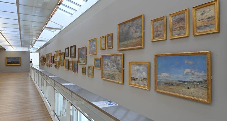 Modern art museum corridor with skylight ceiling and numerous impressionist paintings hung on grey walls.