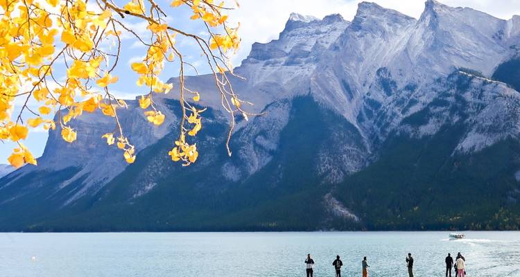 Un tranquilo lago turqueso se encuentra bajo picos grises y escarpados con hojas de otoño enmarcando el primer plano y pequeños visitantes en la orilla.