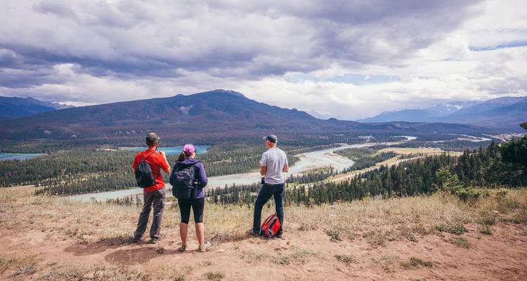 Tres excursionistas con mochilas contemplan desde una cresta cubierta de hierba un valle fluvial sinuoso respaldado por montañas boscosas.