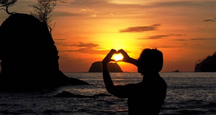 Persona haciendo un gesto de corazón con las manos durante un atardecer en la playa.