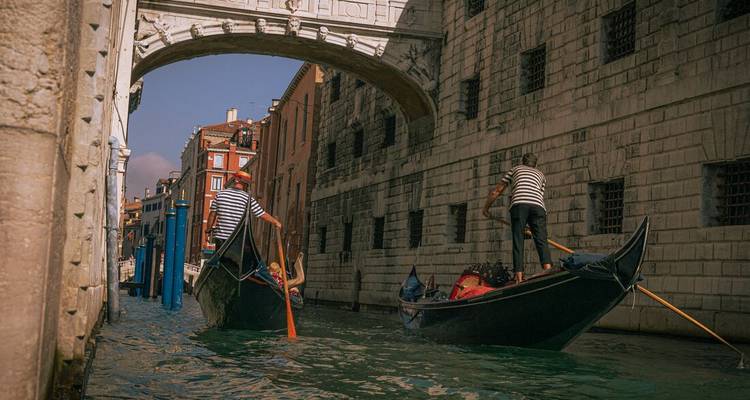 Deux gondoliers en chemises rayées rament leurs gondoles sous un pont de pierre le long d'un canal vénitien étroit.