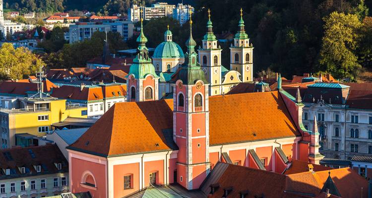 Aerial cityscape of Ljubljana with the pink Franciscan Church and green-domed towers set against forested hills.