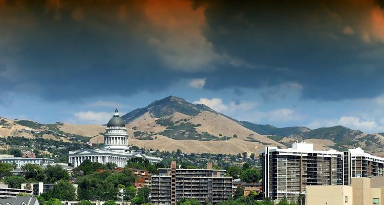 Skyline of Salt Lake City with Utah State Capitol and mountains beneath dramatic storm clouds.