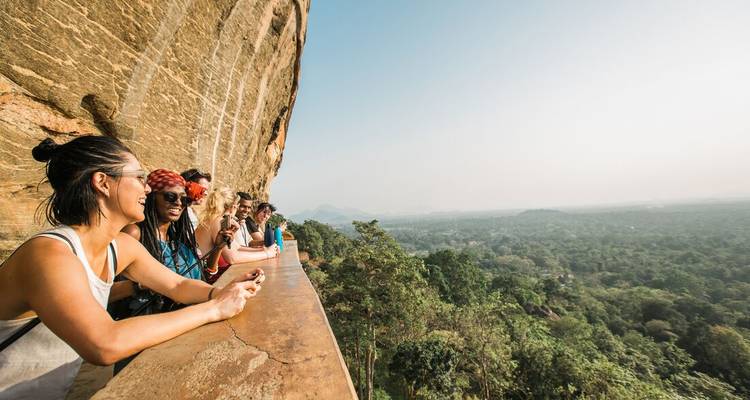 Travellers lean over the ancient stone parapet of Sigiriya Rock, admiring a vast forest plain below in hazy morning light.