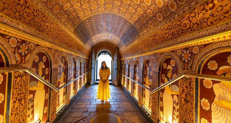 A woman in a flowing yellow dress stands in an ornately painted Buddhist corridor lit by warm spotlights.
