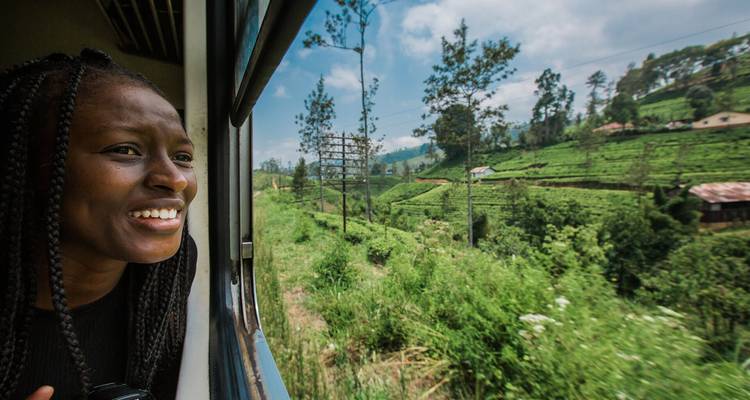Una mujer sonríe desde la ventana abierta del tren pasando junto a exuberantes plantaciones de té verde bajo un cielo azul.