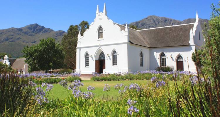 Weiße Kirche mit Gärten vor einer Bergkulisse.