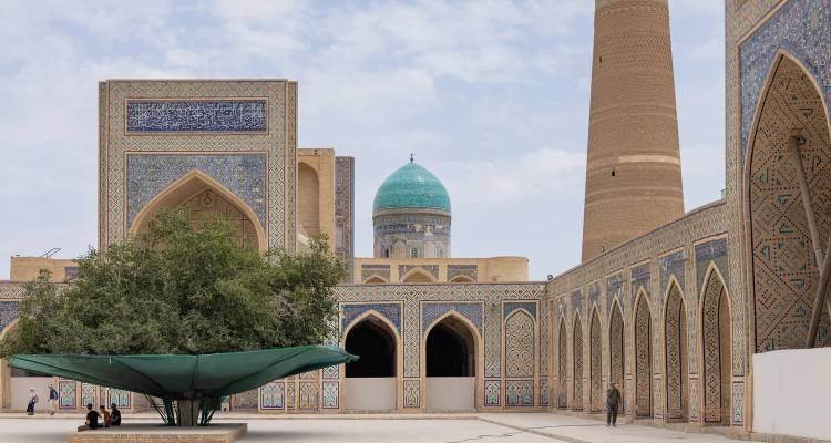 Patio de una madrasa ornamentada en Bujará con arcos de azulejos, una cúpula turquesa y un minarete elevado bajo un cielo parcialmente nublado