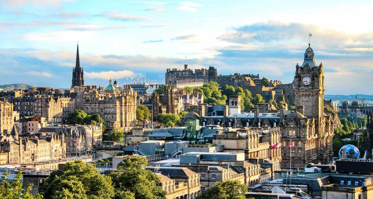 Weitläufige Skyline von Edinburgh mit dem Balmoral Uhrenturm und der Burg vor einem teilweise bewölkten Abendhimmel.