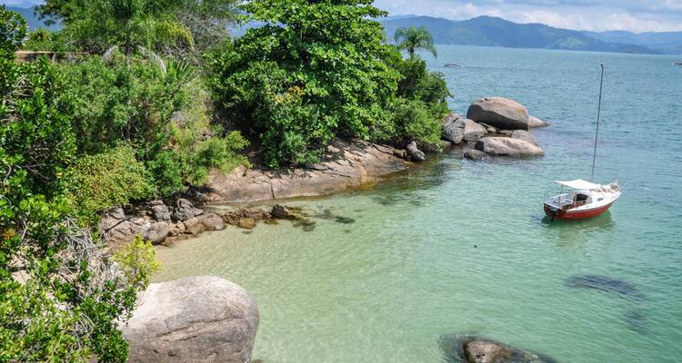 Pequeño velero blanco anclado en una tranquila cala de aguas turquesas rodeada de rocas exuberantes y vegetación.