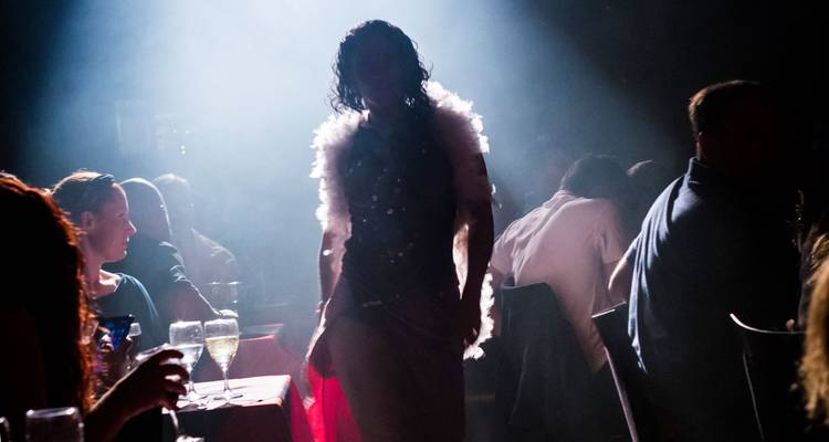 Escena de cabaret con poca luz con una artista en boa de plumas silueteada por un reflector entre la audiencia sentada con bebidas.