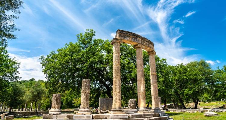 Les anciennes colonnes du Philippeion entourées d'arbres verts luxuriants sous un ciel bleu strié
