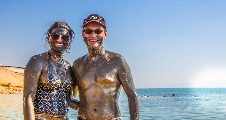 Stel bedekt met modder glimlachend bij de zee onder een heldere hemel.