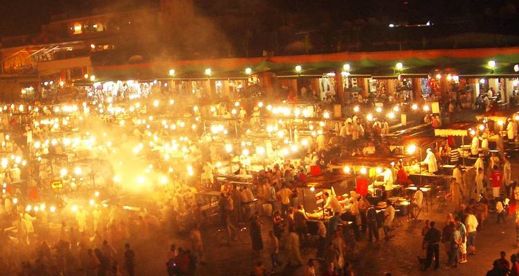 Escena nocturna de la Jemaa el-Fnaa llena de comensales y puestos de comida iluminados, con humo y luces creando una atmósfera festiva y brumosa.