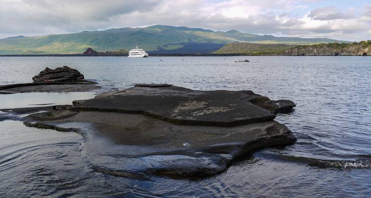 Bahía tranquila con un yate anclado en alta mar, una plataforma de roca de lava plana en primer plano y colinas volcánicas verdes más allá.