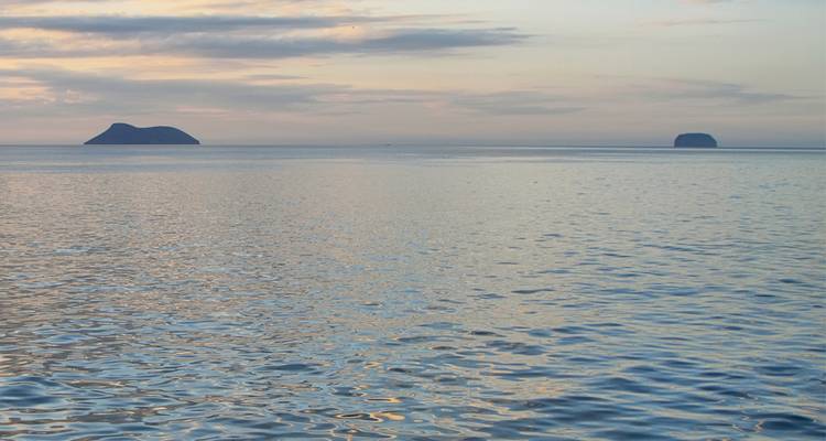 Amplio tramo de océano tranquilo al atardecer con dos pequeñas islas silueteadas en el horizonte bajo cielos de tonos pastel.