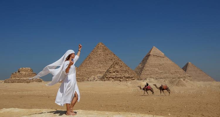 Vrouw in een wapperende witte jurk viert feest voor het trio van de Giza-piramides met kamelen in de verte.