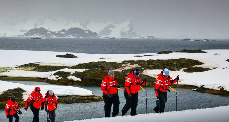 Kleine trekking groep in rode parkas loopt over besneeuwde Antarctische terrein met rotsige eilandjes en zee erachter
