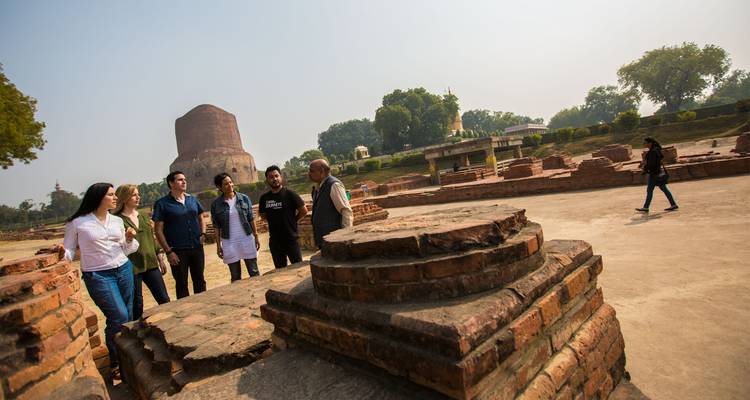 Un pequeño grupo de viajeros escuchando a un guía entre ruinas de ladrillo con la Estupa Dhamek cilíndrica dominando el fondo.