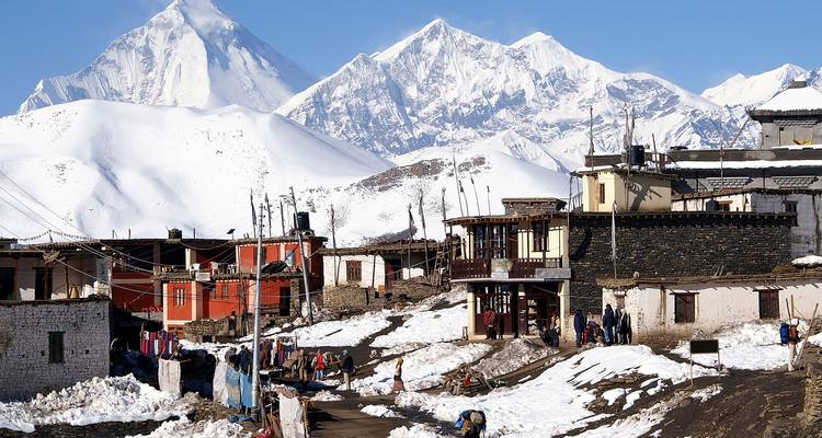 Pueblo himalayo nevado con casas coloridas y enormes picos blancos al fondo.