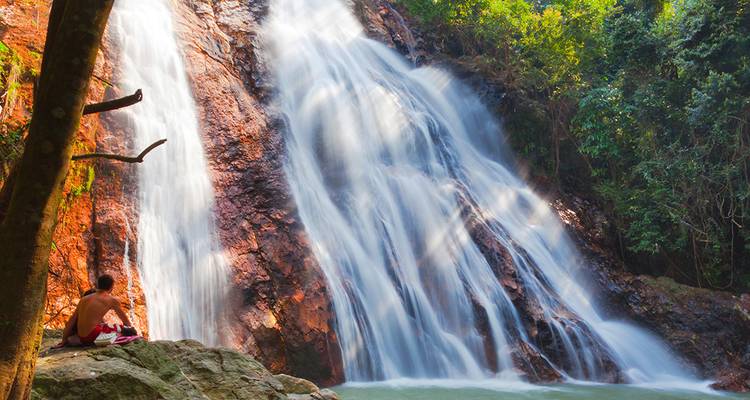 Sanfter verschwommener Wasserfall, der über rotes Gestein herabfließt, während eine Person auf einem Stein sitzt und die Szene bewundert.