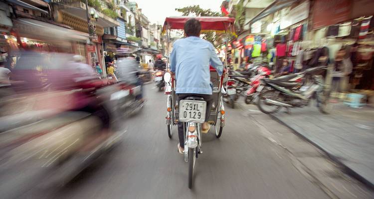 Escena callejera con movimiento desenfocado desde la parte trasera de un ciclo pedaleando por el animado Barrio Antiguo de Hanoi