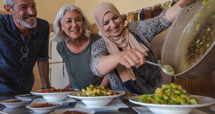 Travellers smile while a local woman pours vegetables into dishes during a cooking lesson.