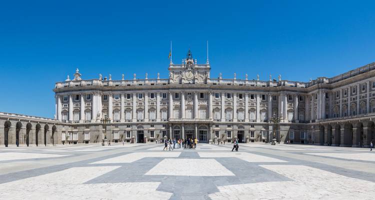 Uitgebreid uitzicht op het Koninklijk Paleis van Madrid en het grote stenen plein eronder onder een heldere blauwe lucht.