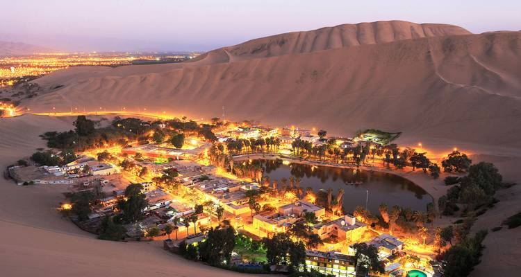 Desert oasis village illuminated at twilight surrounded by towering sand dunes