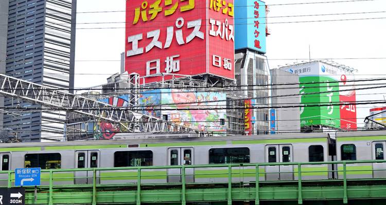 Een groene pendeltrain passeert onder een netwerk van stroomlijnen met felle anime-billboards die hoog boven uittorenend.