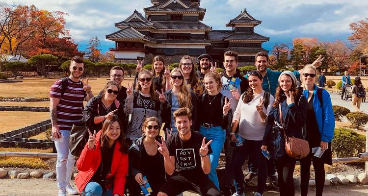 Un grand groupe touristique pose en faisant des signes de paix devant un château japonais historique lors d'une journée d'automne.