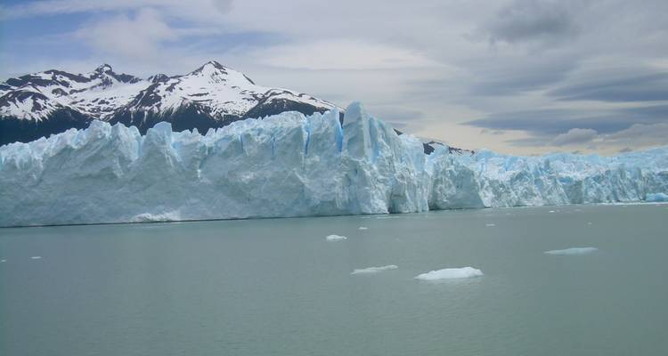 Massive blaue Eismauer des Perito-Moreno-Gletschers, die sich über ruhigem graugrünem Gletscherwasser mit schneebedeckten Gipfeln dahinter erhebt