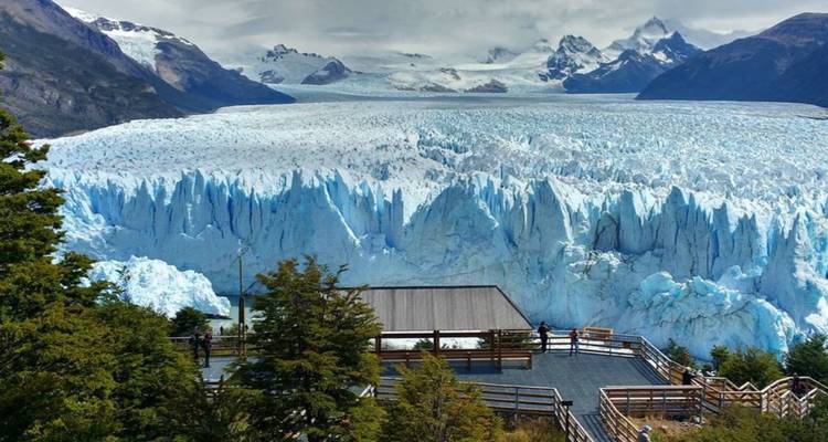 Besucher stehen auf einer hölzernen Terrasse und blicken auf die ausgedehnte, von Spalten durchzogene Oberfläche des Gletschers Perito Moreno, die sich bis zum Horizont erstreckt