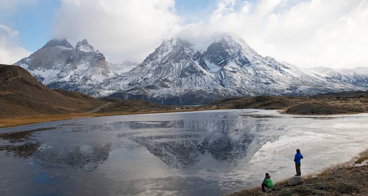 Schneebedecktes Massiv, das sich in einer dünnen Eisschicht auf einem Bergsee in großer Höhe spiegelt, während zwei Personen über das Wasser blicken