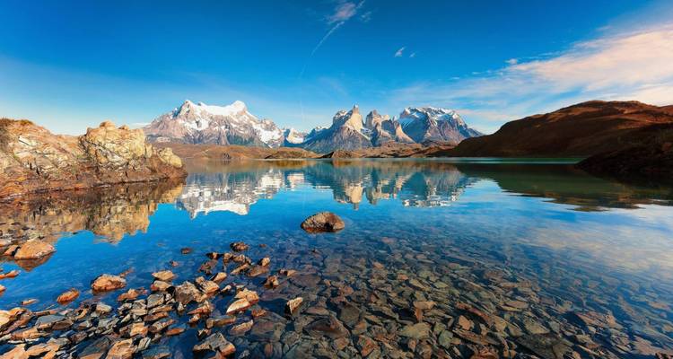 Kristallklarer See mit felsiger Küste, der die dramatischen Türme der Torres del Paine unter einem leuchtend blauen Himmel spiegeln