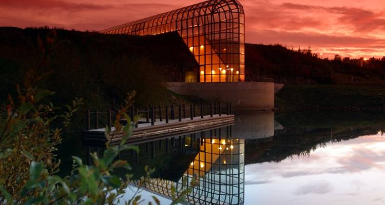 Edificio moderno de cristal reflejándose en agua tranquila al atardecer.