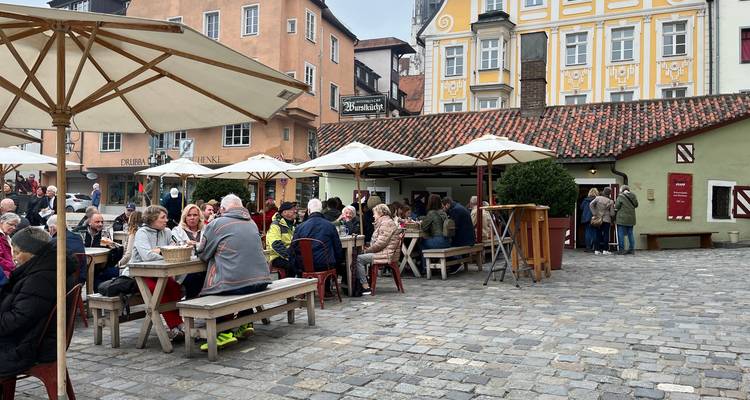 Des foules savourent nourriture et boissons à des tables extérieures sous de grands parasols sur une place bavaroise pavée de galets.