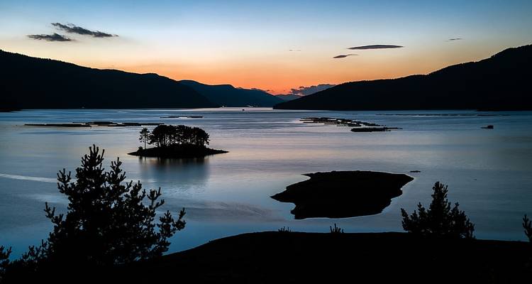 Coucher de soleil sur un lac avec des îles bordées d'arbres et des silhouettes de montagnes.
