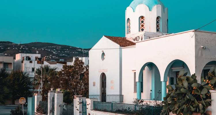 Witgekalkte Grieks-orthodoxe kerk met blauwe koepel en bogen tegen een helderblauwe lucht.