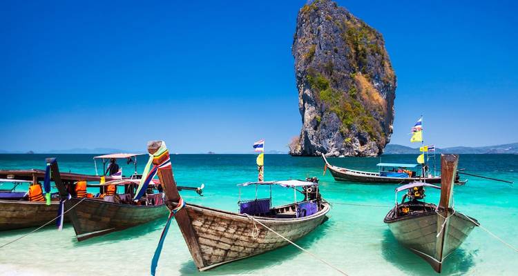 Klassische thailändische Longtail-Boote, die in kristallklarem türkisfarbenem Meer vor einer steilen Kalksteinnadel treiben.