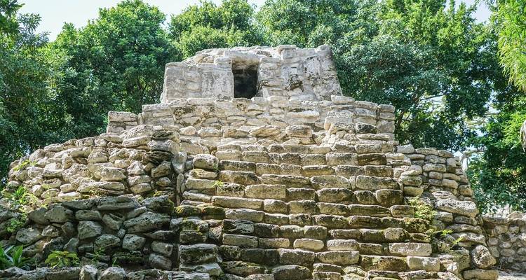 Small stepped stone pyramid structure surrounded by lush jungle trees under a bright blue sky.