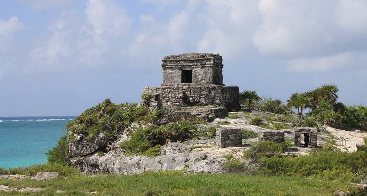 Small stone Mayan ruin perched on a rocky cliff above bright Caribbean waters and a turquoise sea.
