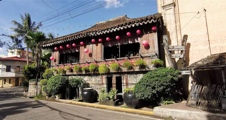 Casa de madera histórica adornada con farolillos rosas y plantas en macetas a lo largo de una calle tranquila.