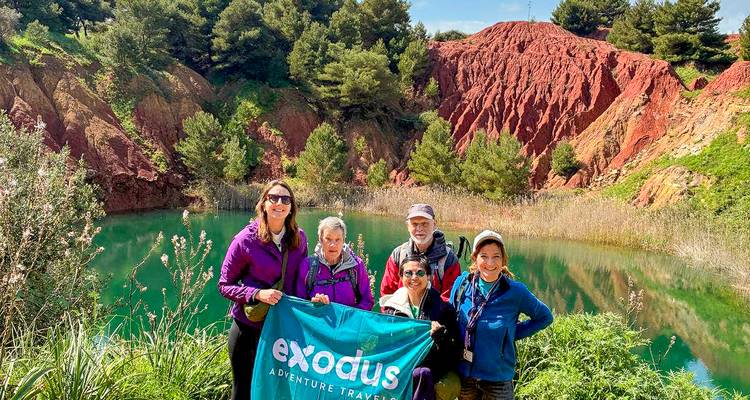 Cinco excursionistas sosteniendo un banner de Exodus Adventure Travels se encuentran junto a un lago de un verde esmeralda vivido rodeado de acantilados rojos impresionantes y vegetación.