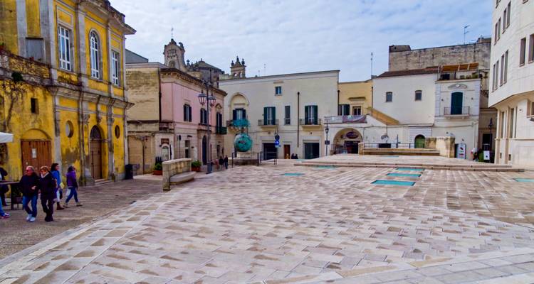 Una tranquila plaza soleada rodeada de edificios históricos en colores pastel, puertas arqueadas y campanarios en un pueblo del sur de Italia.