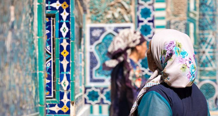 Frauen in farbenfrohen Kopftüchern bewundern die aufwendige blau- und türkisfarbene Fliesenarbeit in einem zentralasiatischen Mausoleum.