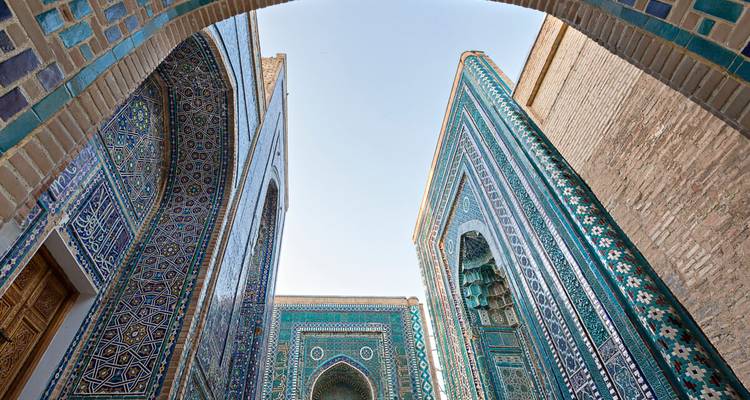 Der Blick nach oben zwischen den aufragenden, türkisfarben gekachelten Mausoleuumswänden in Richtung eines blassen Himmels in Samarkands Shah-i-Zinda-Komplex.