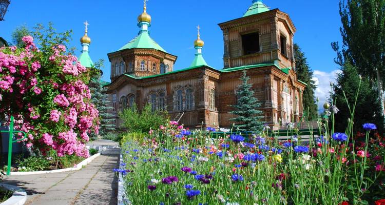 Eine hölzerne russisch-orthodoxe Kathedrale mit grünen Dächern und goldenen Kuppeln erhebt sich hinter leuchtenden Sommerblumenbeeten.